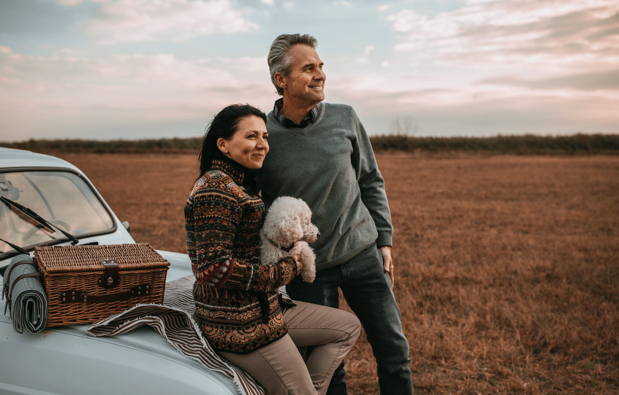 Smiling mature couple with their small dog leaning against a vintage car in a field